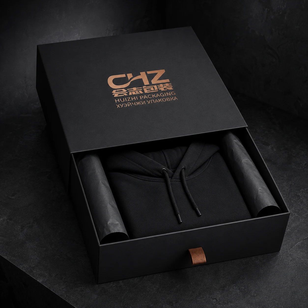 Partly open matte black rigid drawer box holding a folded black hoodie between rolled black tissue supports.