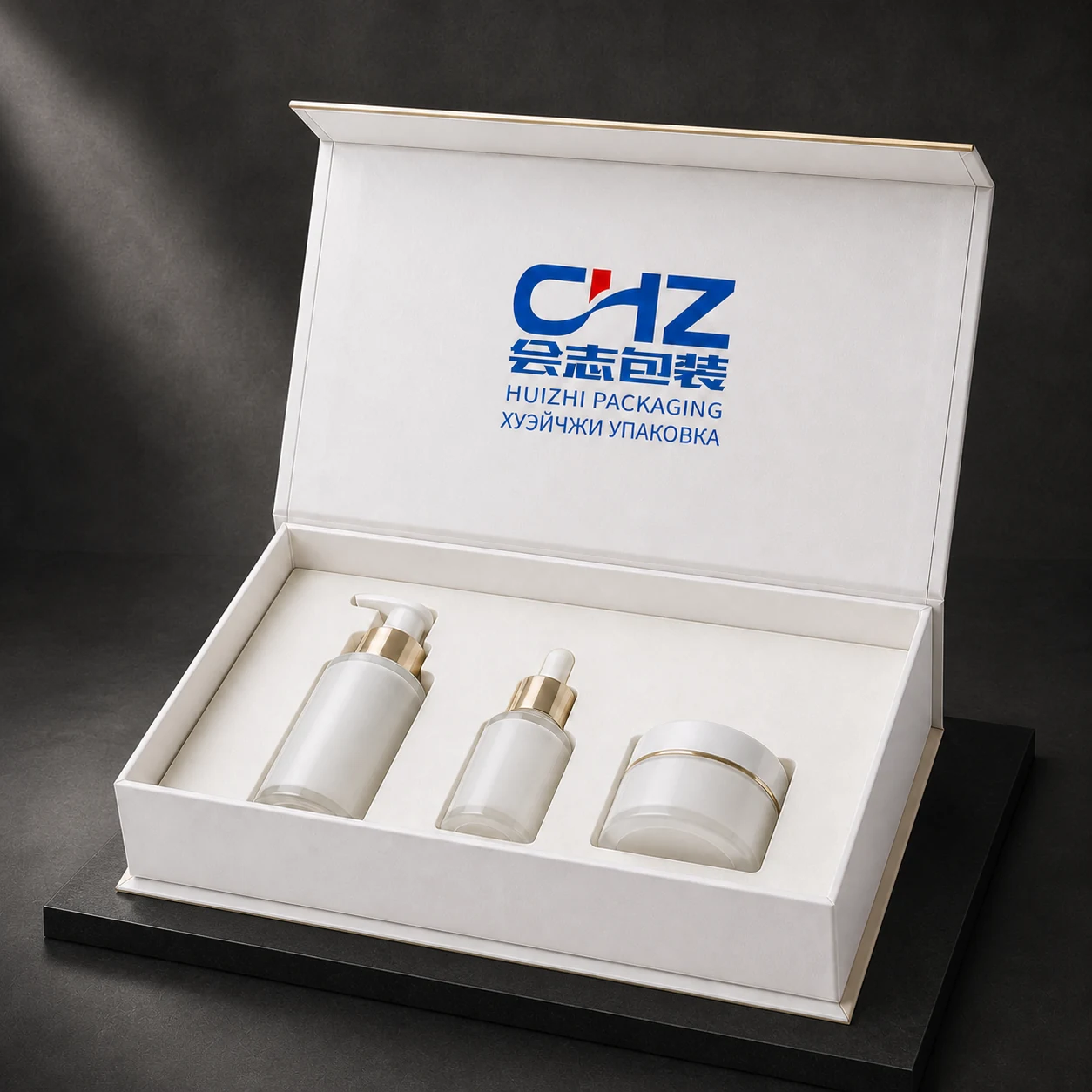 Open pearl ivory rigid skincare gift box holding a pump bottle, dropper bottle, and cream jar on a black stone platform.