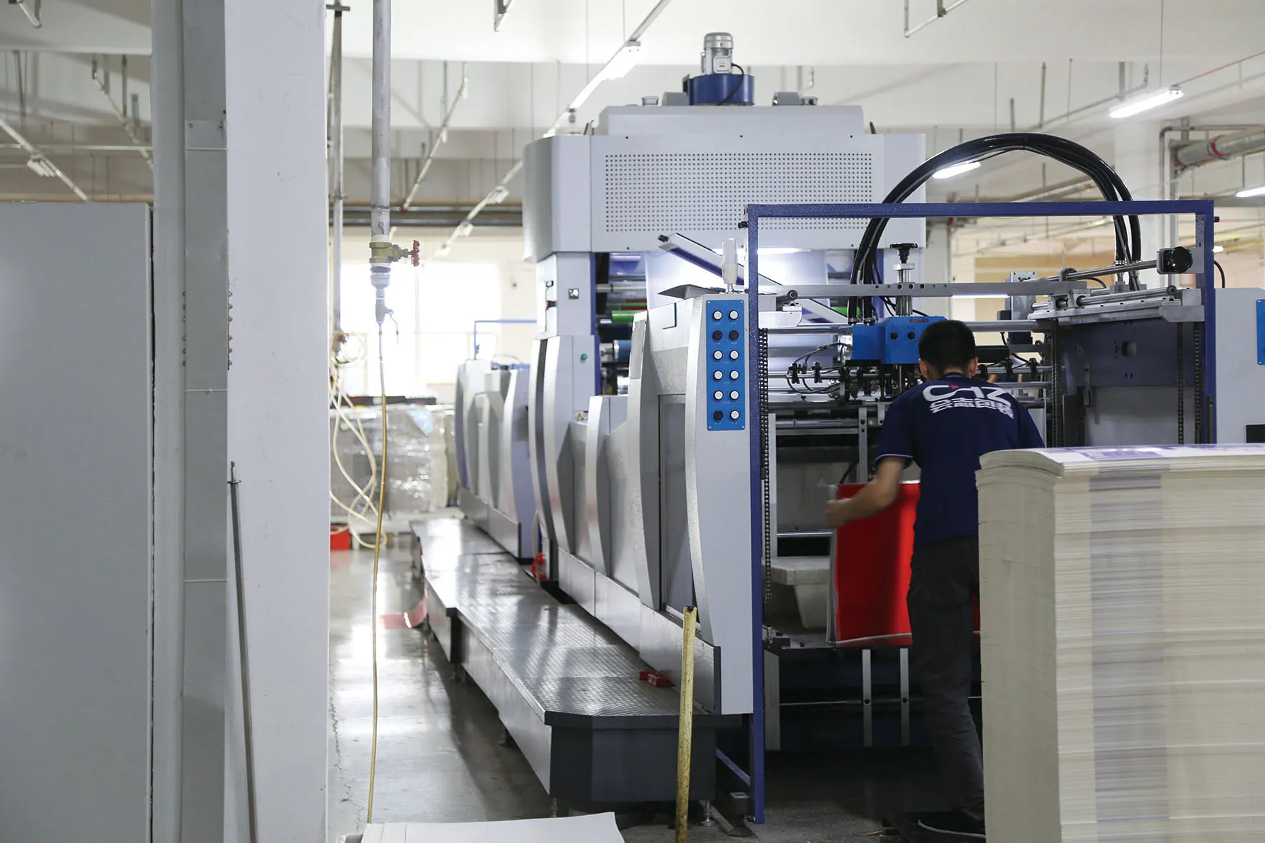 An operator working beside production equipment and a stack of paper sheets