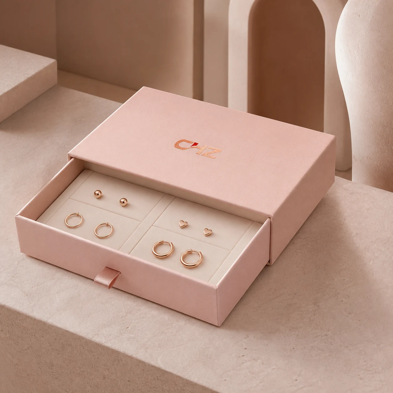 Open blush pink slim jewelry drawer box displaying four pairs of simple unbranded earrings in a fitted tray.